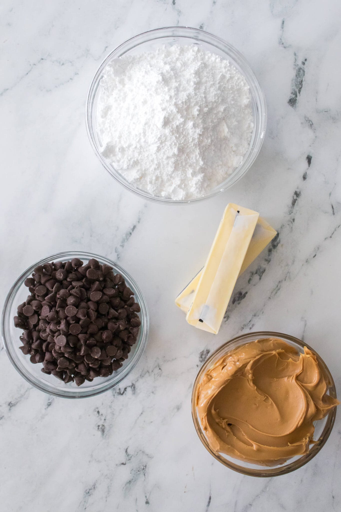 Ingredients on a marble surface include powdered sugar, chocolate chips, two sticks of butter, and a bowl of peanut butter.
