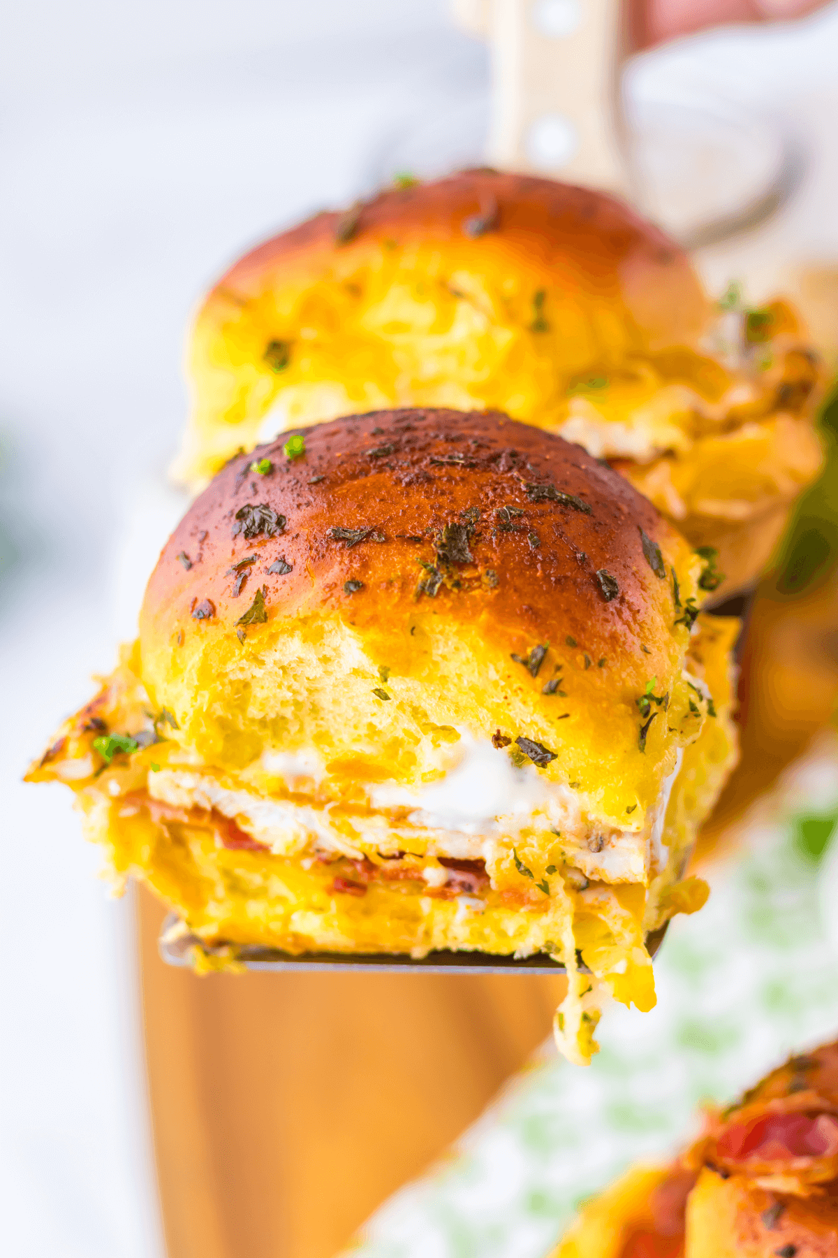 A close-up of a golden-brown Chicken Bacon Ranch Slider filled with gooey melted cheese and herbs, held by a spatula.