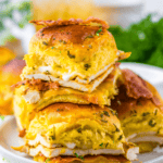 Two stacks of sandwiches with melted cheese on fluffy buns are garnished with herbs and served on a white plate. A green napkin and a drink are in the background.