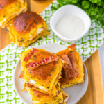 Plate of stacked Chicken Bacon Ranch Sliders on a wooden table, garnished with fresh parsley and served with a bowl of dipping sauce and a glass of soda in the background.