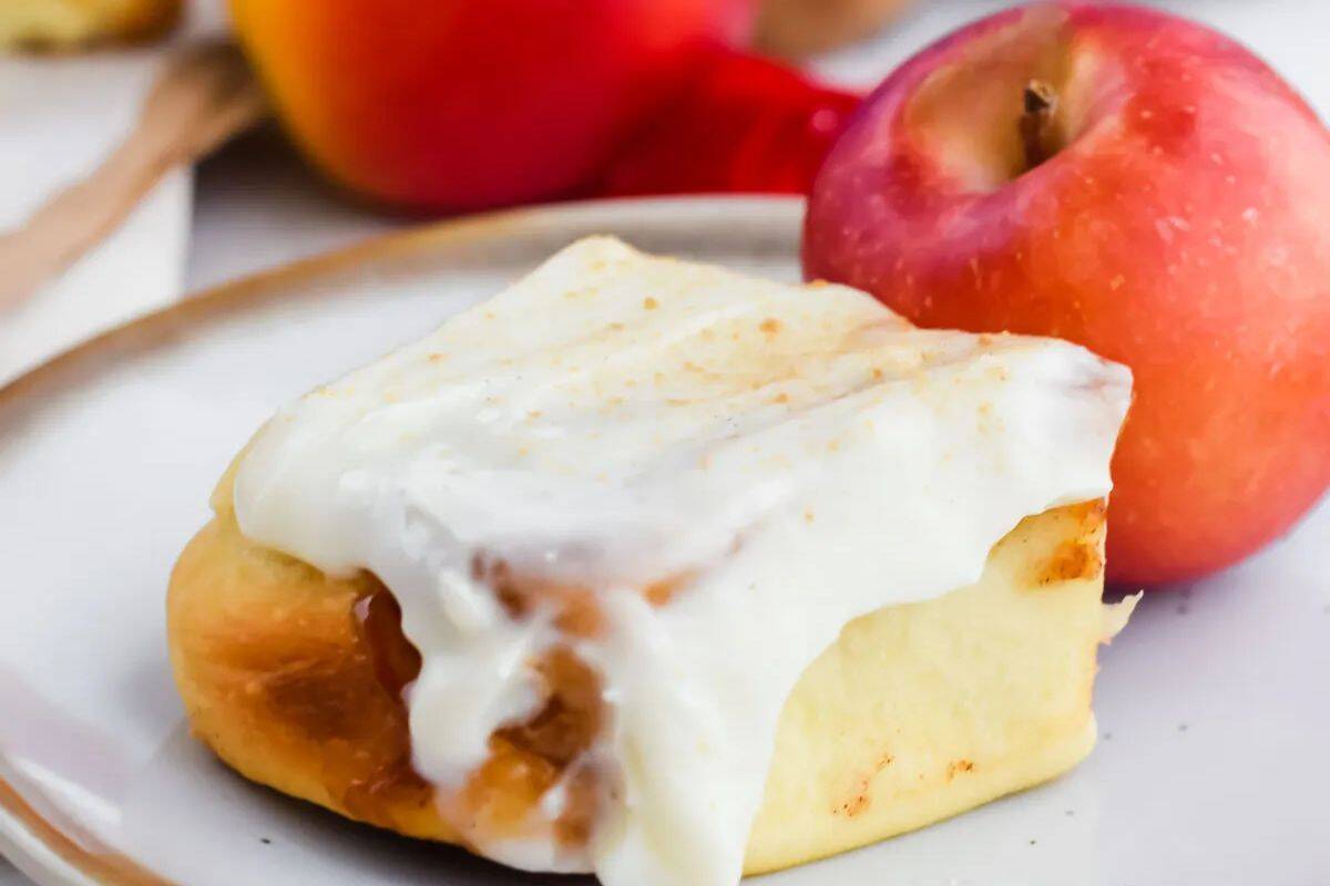 A cinnamon roll with white icing sits on a plate next to a fresh red apple.