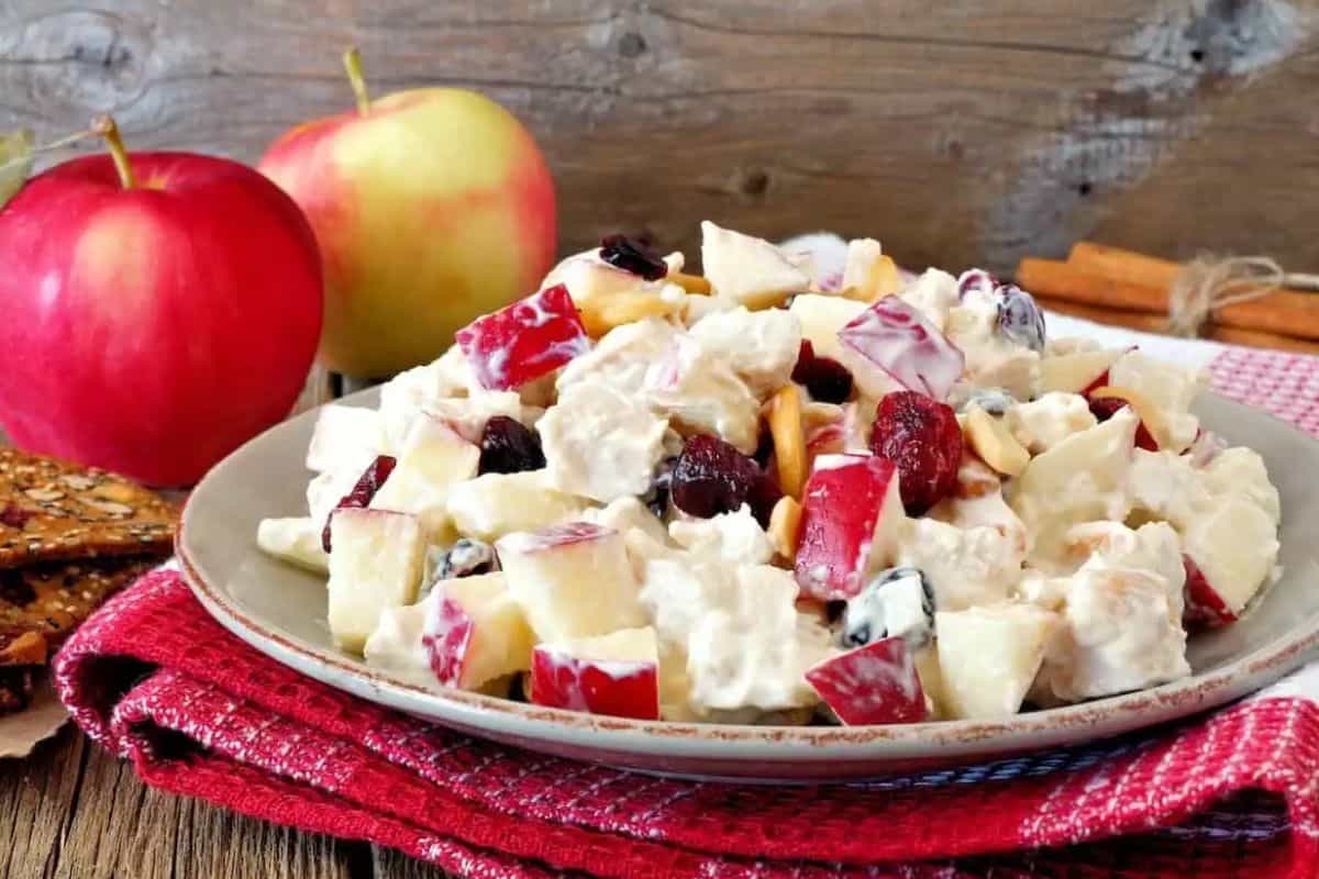 A plate of salad featuring apples and cranberries is displayed on a red cloth, with two apples in the background.