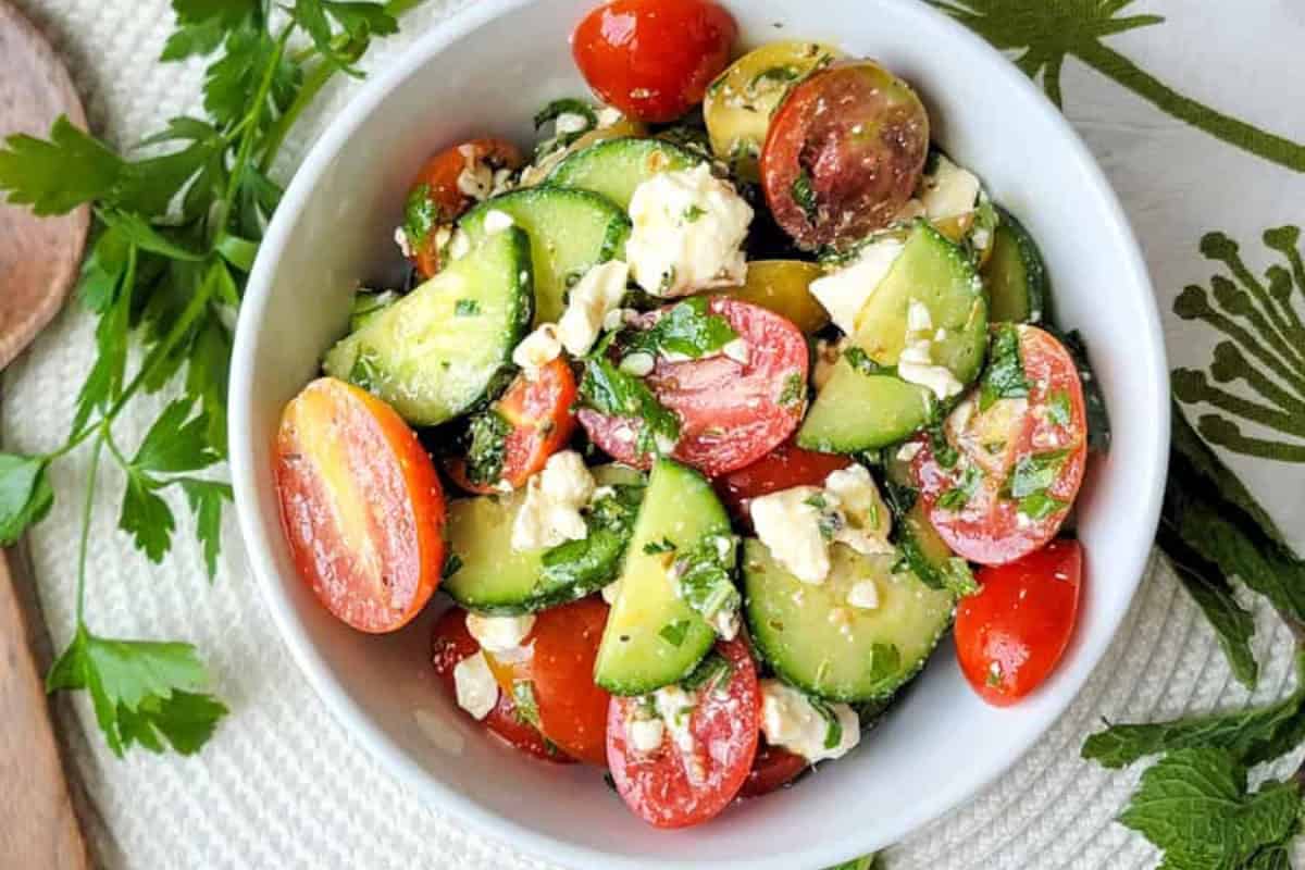 A bowl of salad with sliced cucumbers, cherry tomatoes, crumbled feta cheese, and chopped herbs on a white textured surface—perfect for fresh tomato recipes.