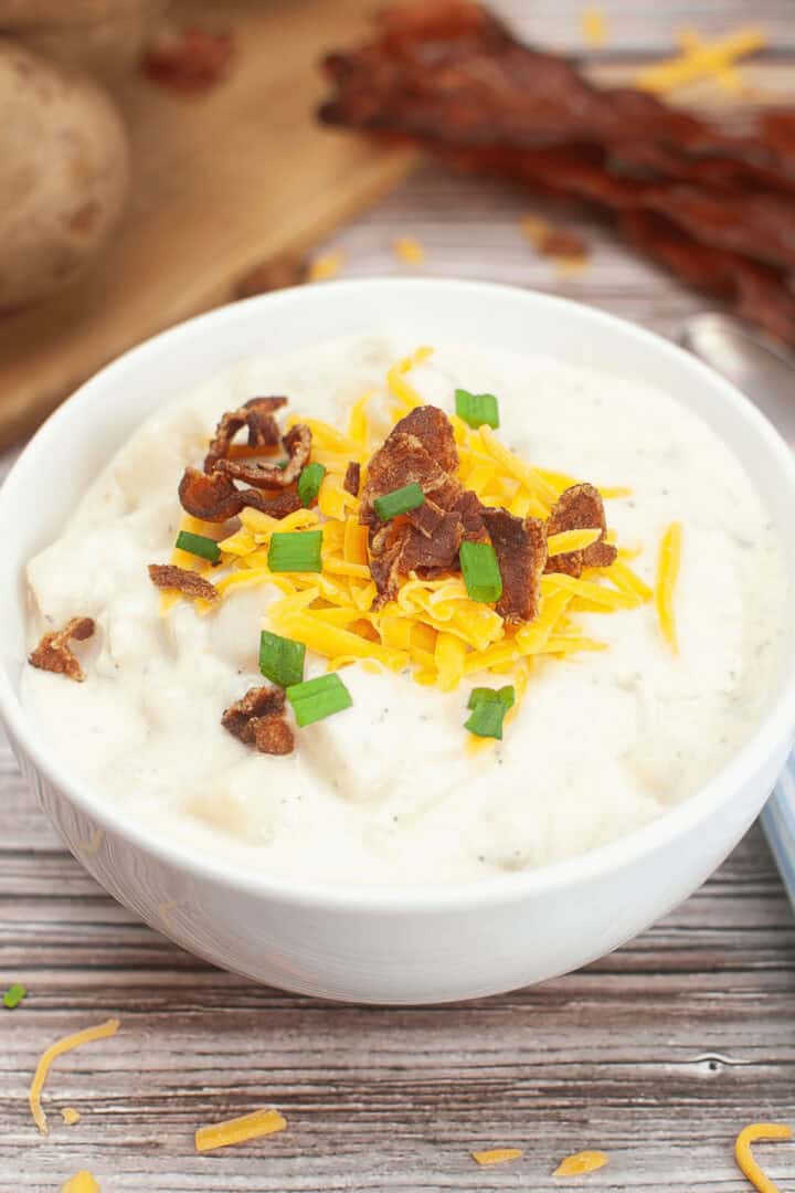 A bowl of creamy potato soup topped with shredded cheddar cheese, crumbled bacon, and chopped green onions, presented as a delectable Crock Pot Potato Soup recipe on a wooden surface.