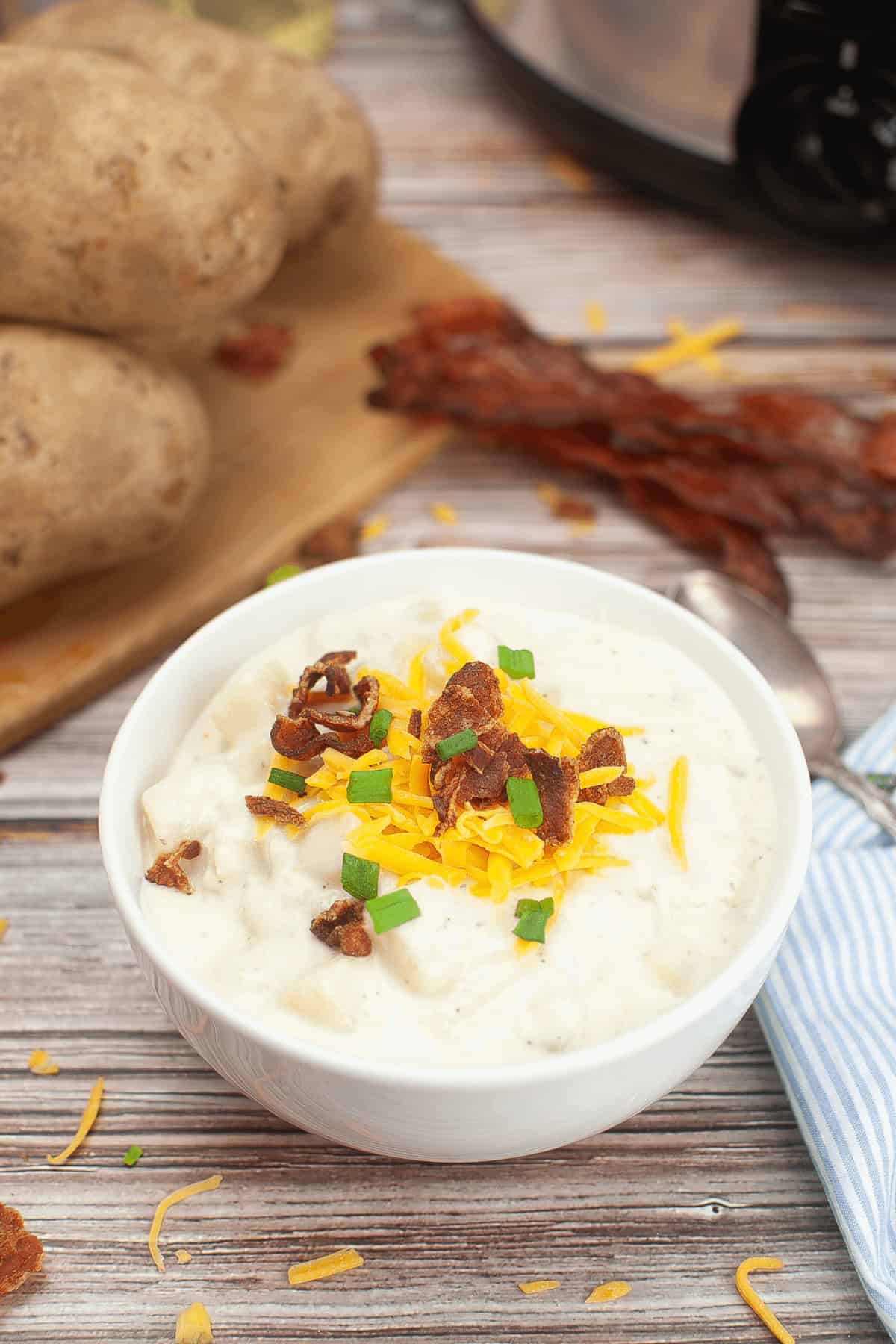 A bowl of Crock Pot Potato Soup Recipe garnished with shredded cheese, bacon bits, and green onions is placed in the foreground. Whole potatoes, a spoon, and a striped napkin are visible in the background.