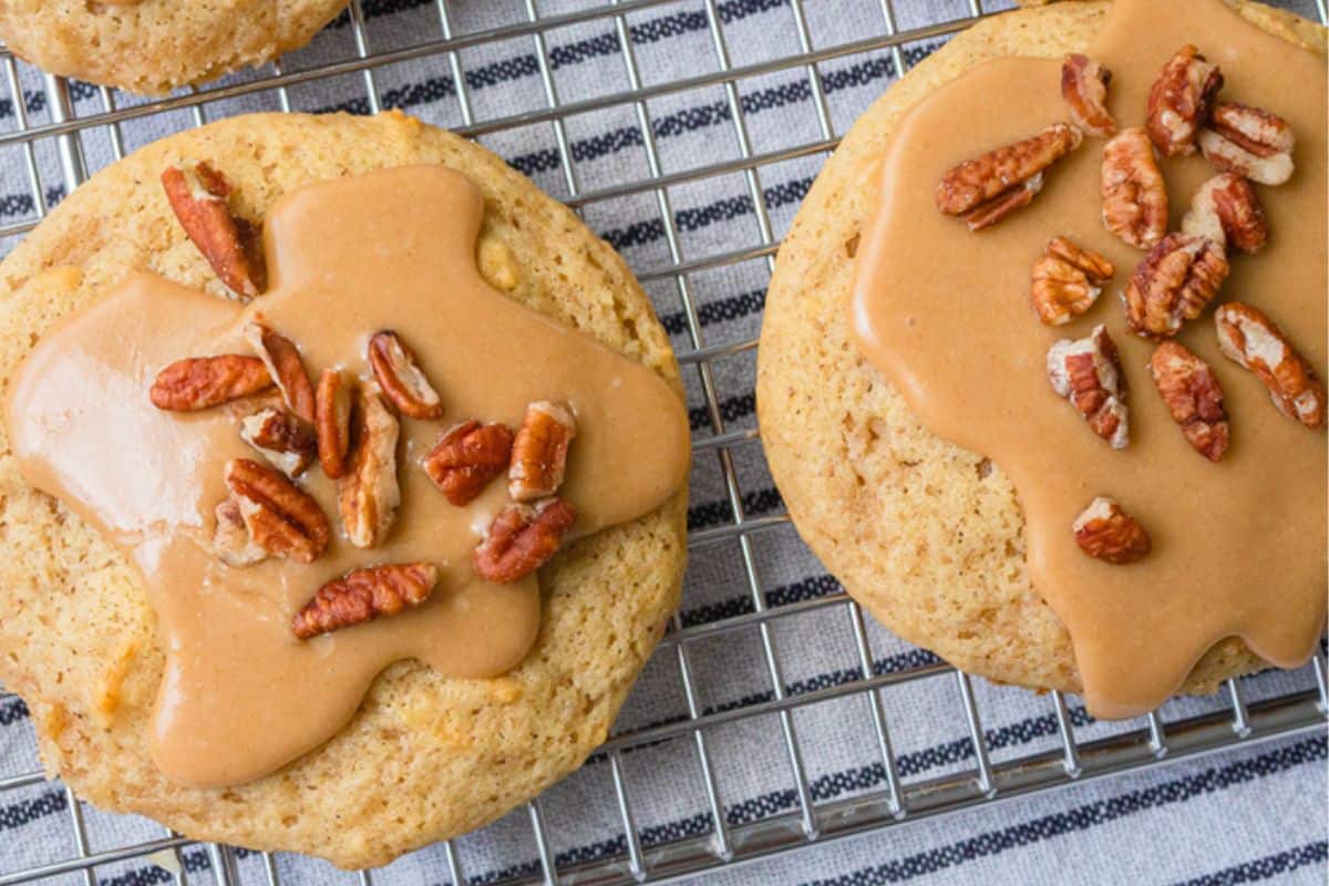 Two apple cider cookies topped with a glossy, runny frosting and pecans rest on a cooling rack, with a striped fabric in the background.