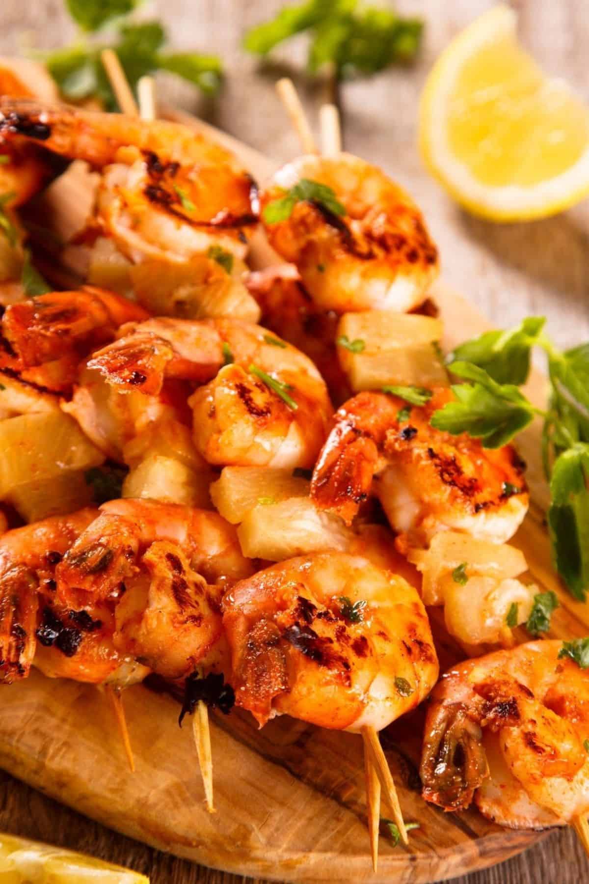 Grilled shrimp skewers with herbs and pineapple pieces, served on a wooden board with lemon wedges and fresh parsley, make for a vibrant and flavorful seafood dish.