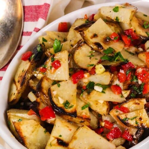 A white dish filled with warm potato salad, featuring grilled potato slices mixed with chopped red peppers, onions, and herbs, placed next to a striped kitchen towel and a metal serving spoon.