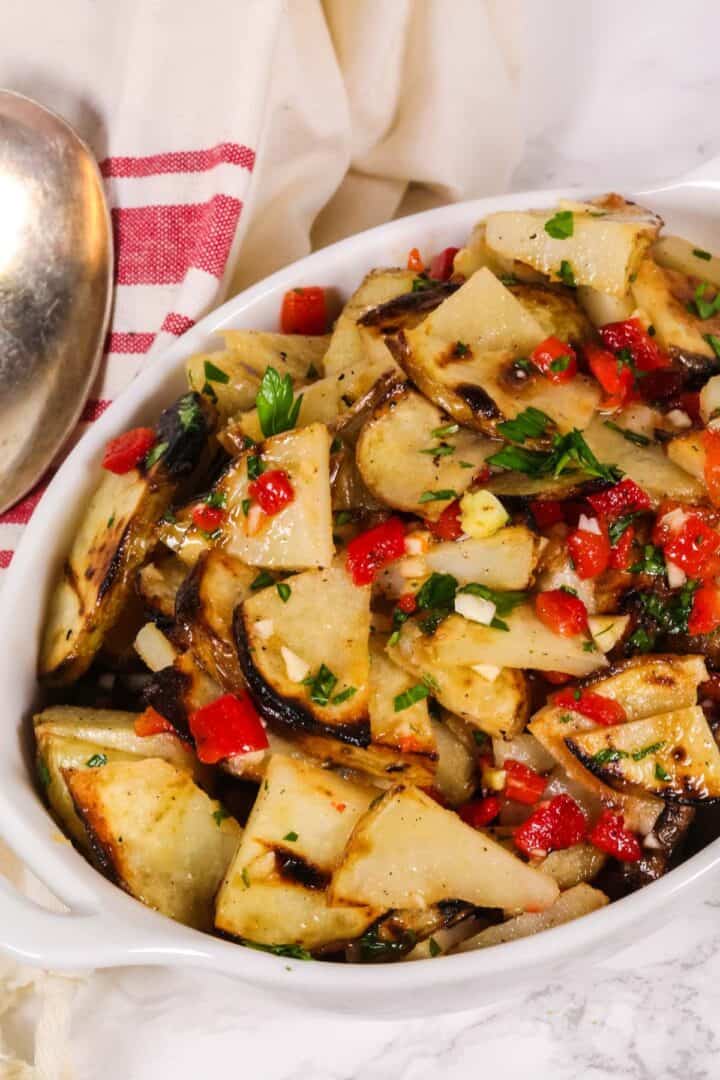 A white dish filled with warm potato salad, featuring grilled potato slices mixed with chopped red peppers, onions, and herbs, placed next to a striped kitchen towel and a metal serving spoon.