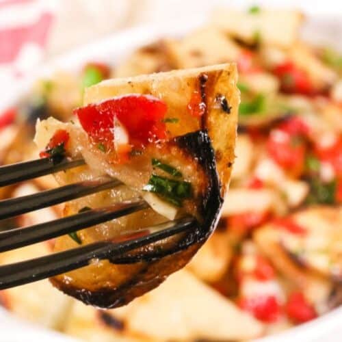 A close-up of a fork holding a piece of grilled chicken topped with chopped tomatoes and herbs, with a bowl of Warm Potato Salad and the same dish in the background.