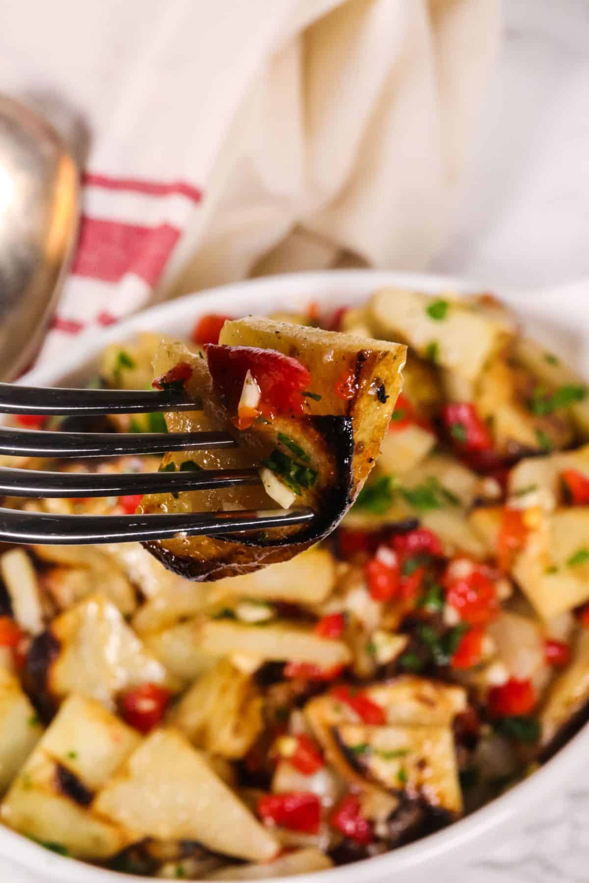 A fork holds a bite of warm potato salad with roasted potatoes, chopped red peppers, and herbs, with a bowl of the same dish in the background.