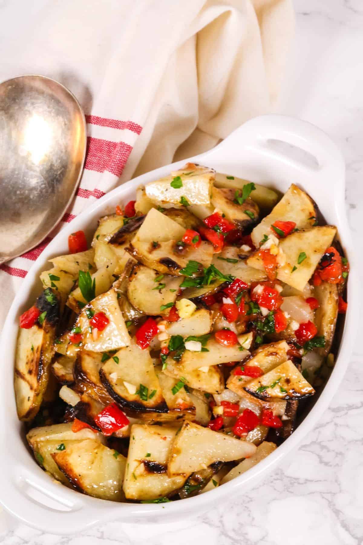 White ceramic dish with roasted potato wedges, chopped red peppers, parsley, and herbs creates a delicious warm potato salad. A silver spoon and white towel with a red stripe are placed nearby on a marble surface.