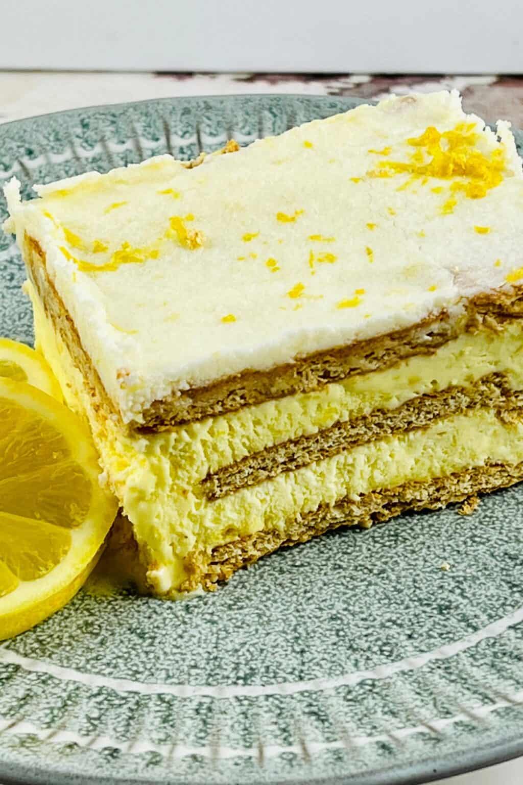 A slice of layered Lemon Icebox Cake with creamy filling and graham crackers, garnished with lemon zest, sits on a green plate next to a lemon slice.