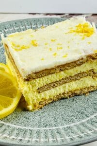 A slice of layered Lemon Icebox Cake with creamy filling and graham crackers, garnished with lemon zest, sits on a green plate next to a lemon slice.