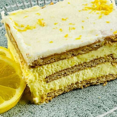 A slice of layered Lemon Icebox Cake with creamy filling and graham crackers, garnished with lemon zest, sits on a green plate next to a lemon slice.