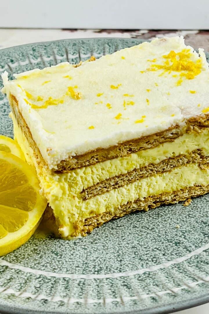 A slice of layered Lemon Icebox Cake with creamy filling and graham crackers, garnished with lemon zest, sits on a green plate next to a lemon slice.