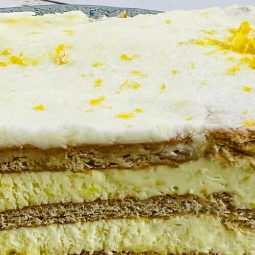 A slice of layered Lemon Icebox Cake with cream and biscuits, garnished with lemon zest, served on a green plate next to lemon slices.