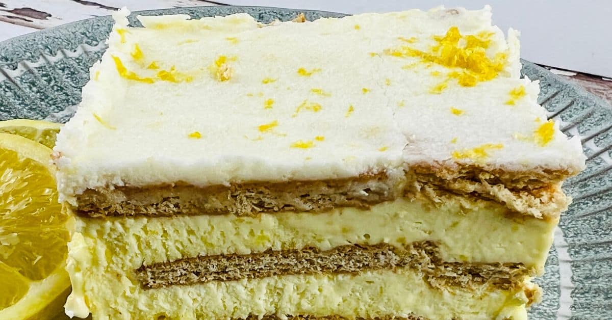 A slice of layered Lemon Icebox Cake with cream and biscuits, garnished with lemon zest, served on a green plate next to lemon slices.