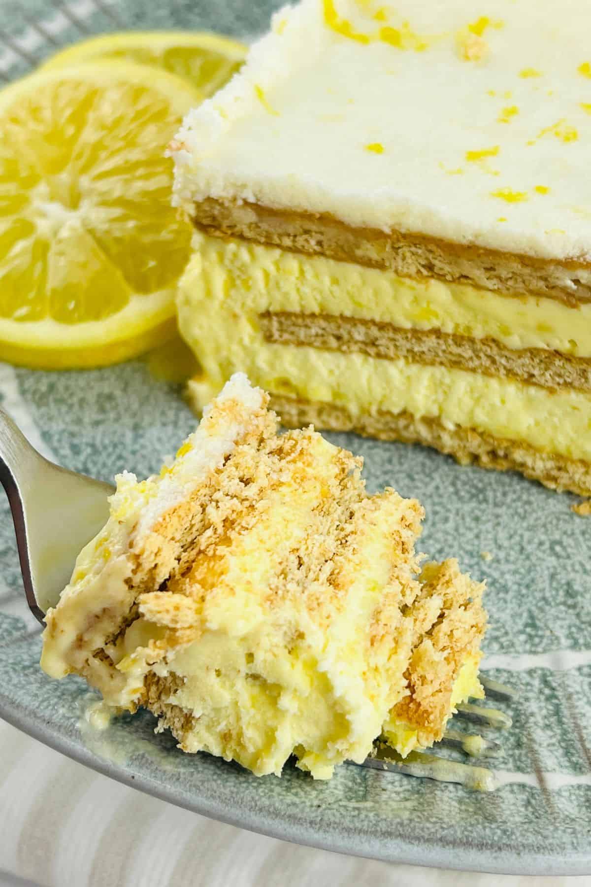 A slice of layered Lemon Icebox Cake with cream and crackers on a plate, garnished with lemon slices; a fork holds a bite-sized piece.