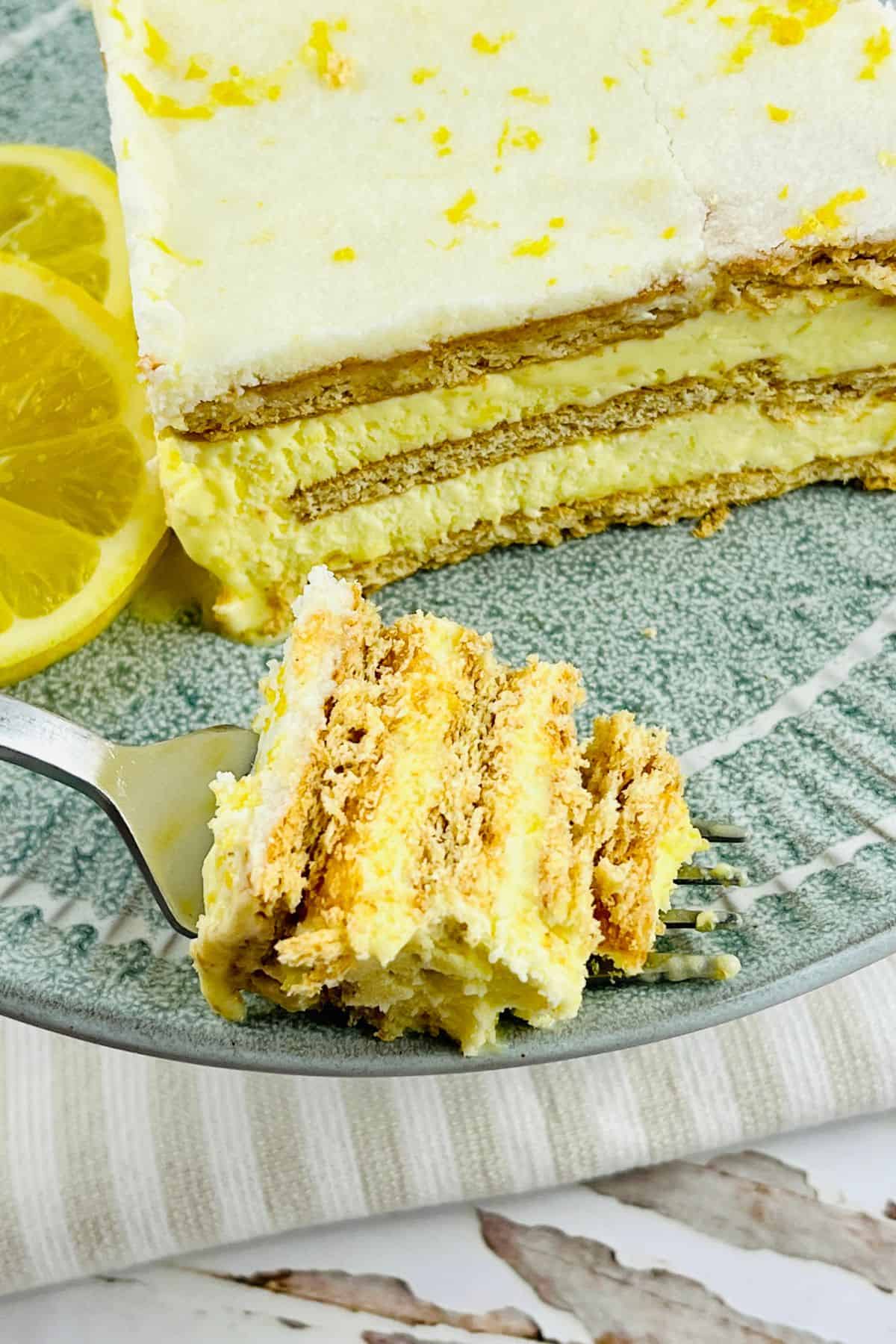 A slice of layered Lemon Icebox Cake with graham crackers and cream sits on a plate, with a forkful in front and lemon slices beside it.