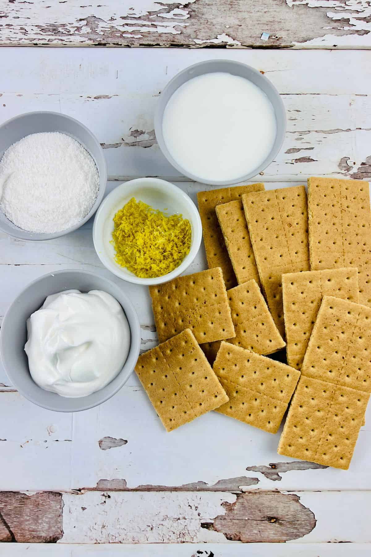 Graham crackers, whipped topping, powdered sugar, milk, and lemon zest are arranged in small bowls on a white rustic wooden surface—everything you need to make a classic Lemon Icebox Cake.