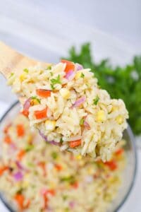 A wooden spoon holds Lemon Orzo Salad—an orzo pasta salad with corn, red bell pepper, red onion, and herbs—above a bowl of the same salad.