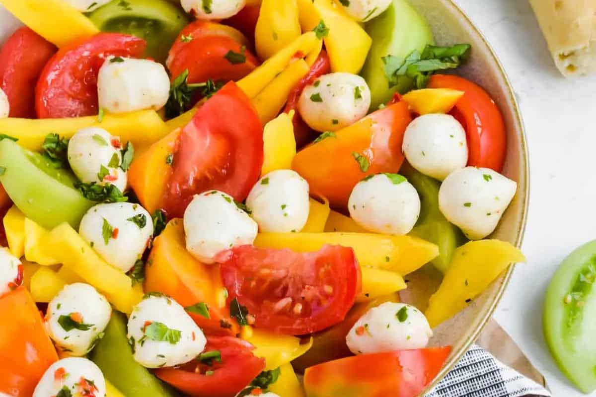 A bowl of salad with fresh tomato—sliced red and yellow tomatoes, mozzarella balls, mango slices, green tomatoes, and chopped herbs—perfect for those who love seasonal recipes.