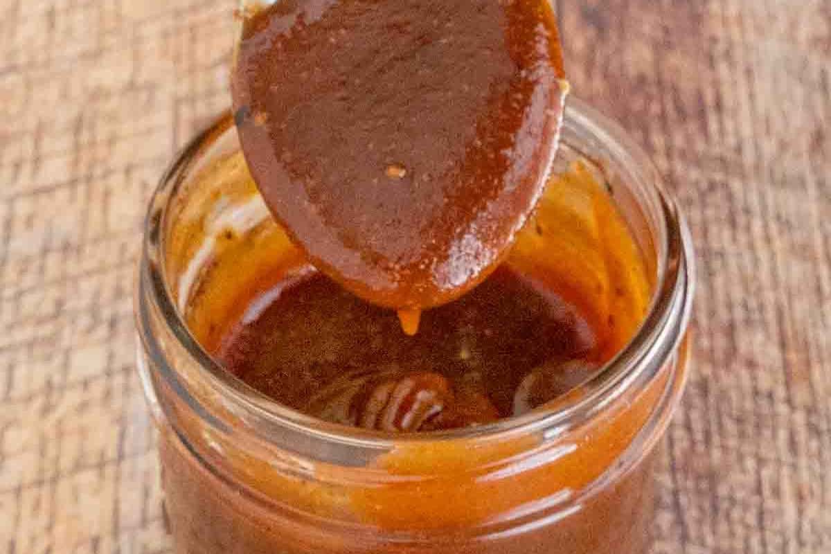 A spoon holding thick brown BBQ sauce hovers above an open glass jar filled with the same sauce, resting on a wooden surface.