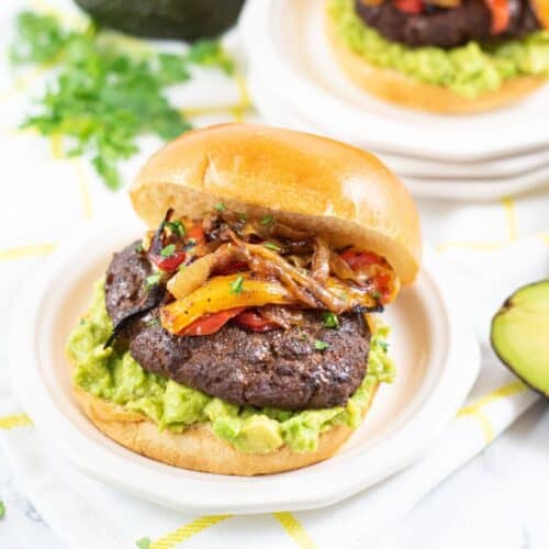 Mexican Hamburger with guacamole, sautéed onions, and bell peppers on a bun, served on a plate. Avocado and cilantro are nearby.