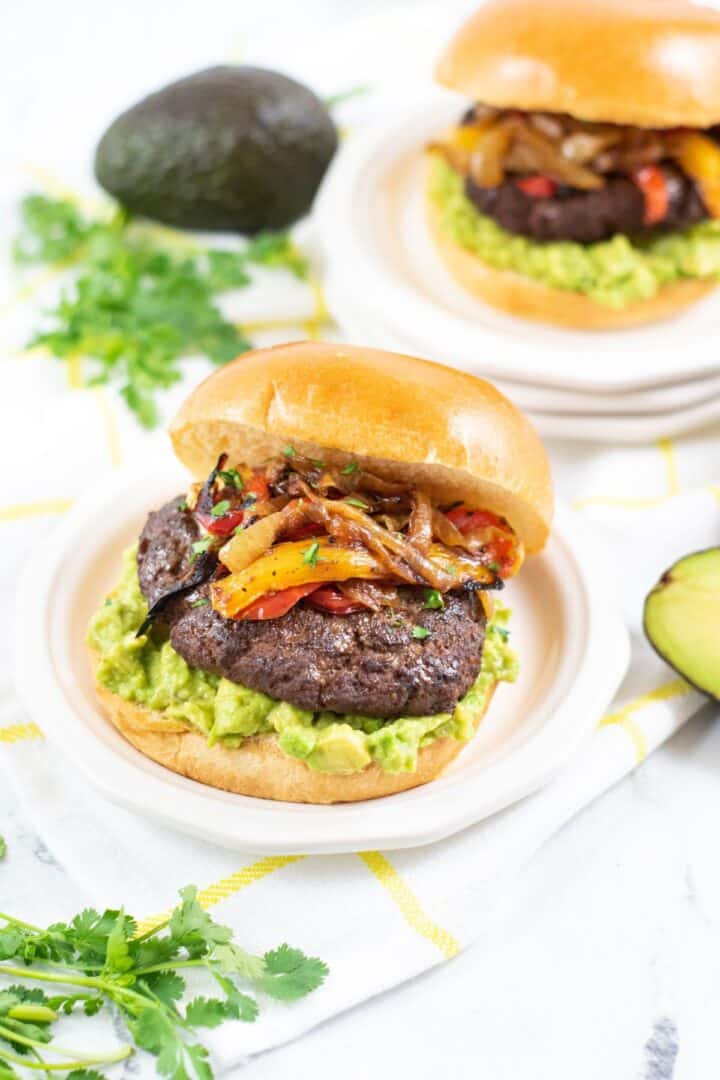 Mexican Hamburger with guacamole, sautéed onions, and bell peppers on a bun, served on a plate. Avocado and cilantro are nearby.