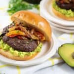 Savor a delicious Mexican Hamburger topped with guacamole, grilled onions, and peppers on a bun, served on a plate with avocado halves and fresh cilantro nearby.