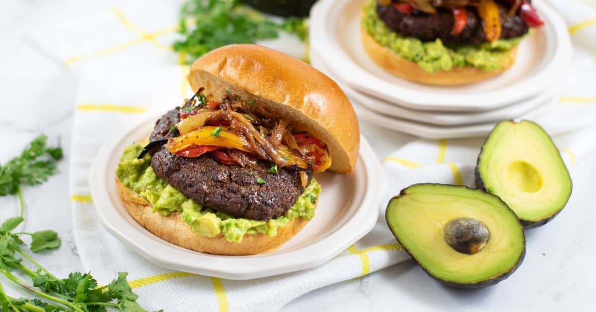 Savor a delicious Mexican Hamburger topped with guacamole, grilled onions, and peppers on a bun, served on a plate with avocado halves and fresh cilantro nearby.