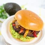 A sandwich on a white plate with guacamole, grilled peppers, onions, and a brioche bun. An avocado and cilantro are in the background.