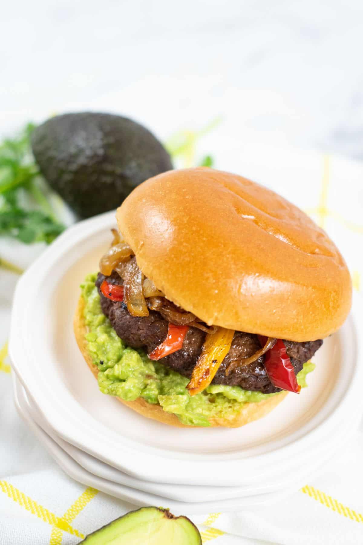 A sandwich on a white plate with guacamole, grilled peppers, onions, and a brioche bun. An avocado and cilantro are in the background.