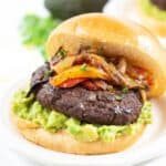 a sandwich with mashed avocado, grilled onions, and peppers on a bun, served on a white plate.