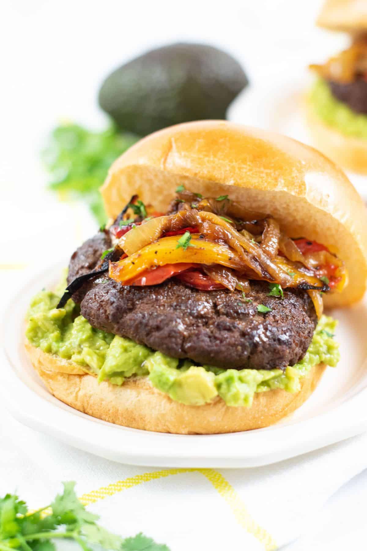 a sandwich with mashed avocado, grilled onions, and peppers on a bun, served on a white plate.