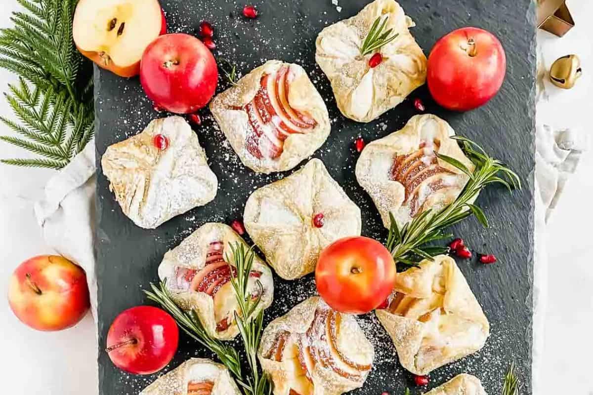 A slate platter displays apple pastries dusted with powdered sugar, red apples, pomegranate seeds, and rosemary sprigs.