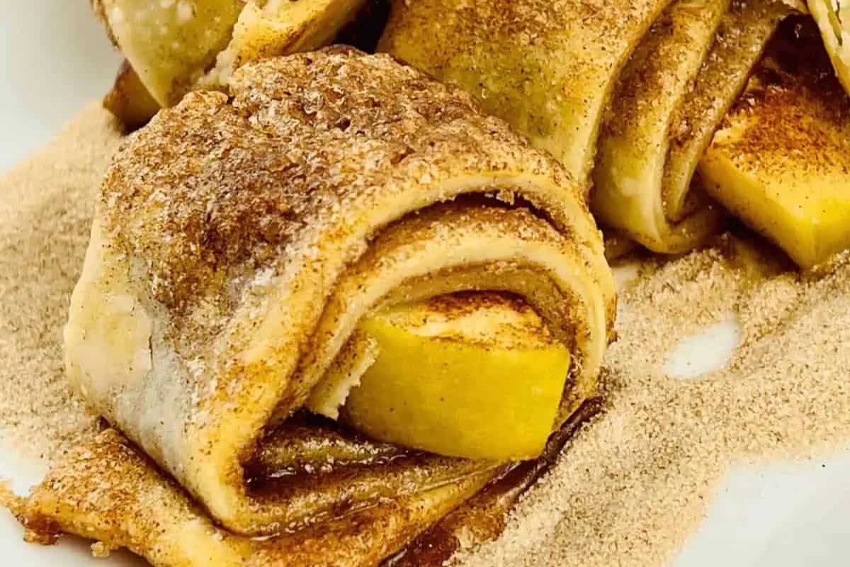 Close-up of pastry dough folded around apple slices, dusted with cinnamon and sugar, resting on a granular surface.