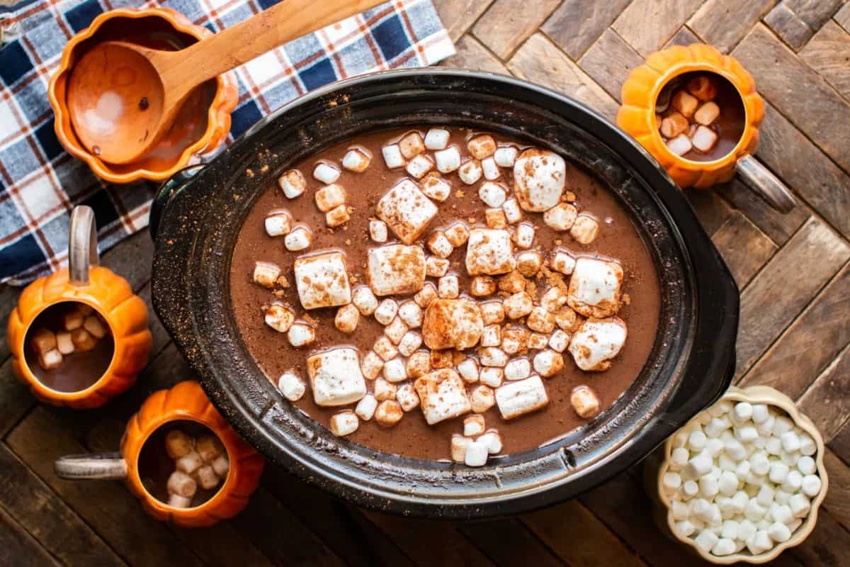 A slow cooker filled with hot chocolate topped with marshmallows, surrounded by mugs, a bowl of mini marshmallows, and a wooden spoon on a checkered cloth—perfect for cozying up with irresistible recipes like pumpkin spice treats.