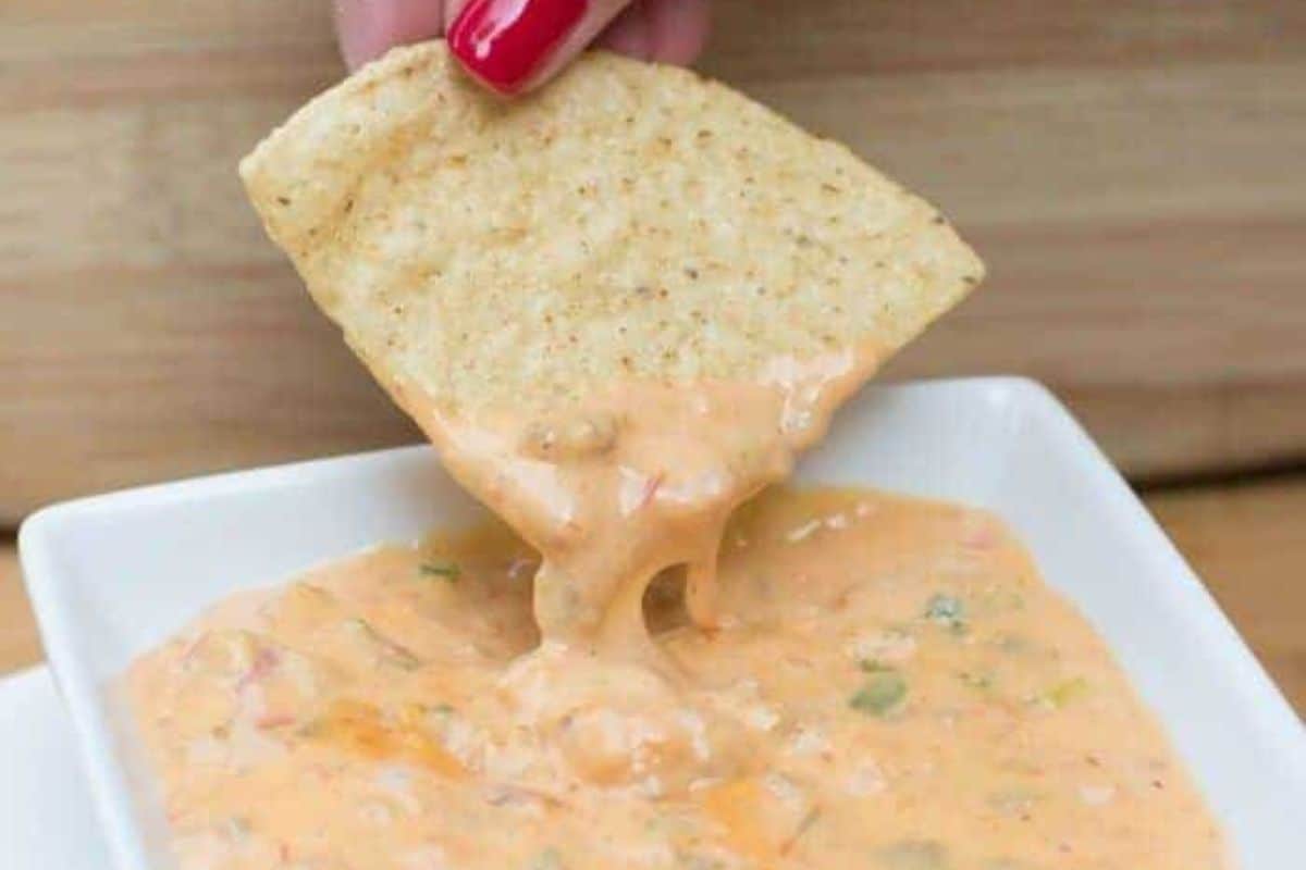 A hand holding a tortilla chip being dipped into a bowl of nacho cheese dip.