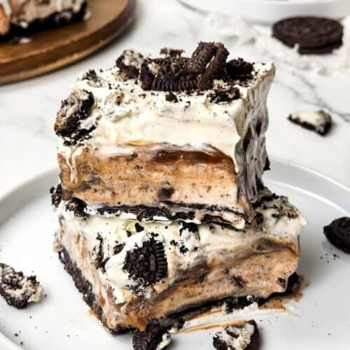 A decadent Oreo Ice Cream Cake features layers of cookies, cream, and caramel stacked on a white plate, finished with a generous topping of crumbled chocolate cookies.