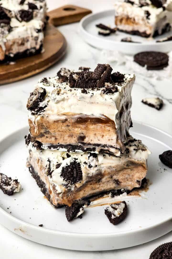 A decadent Oreo Ice Cream Cake features layers of cookies, cream, and caramel stacked on a white plate, finished with a generous topping of crumbled chocolate cookies.