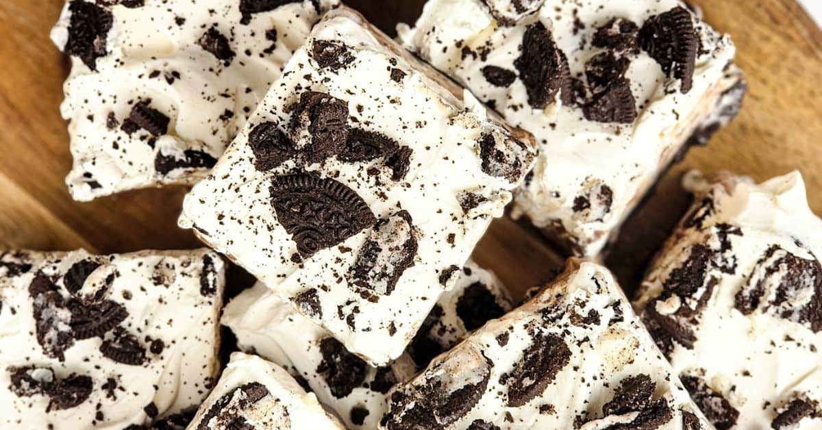 Squares of dessert bars topped with white frosting and crumbled chocolate cookies, reminiscent of an Oreo Ice Cream Cake, are arranged on a wooden surface.