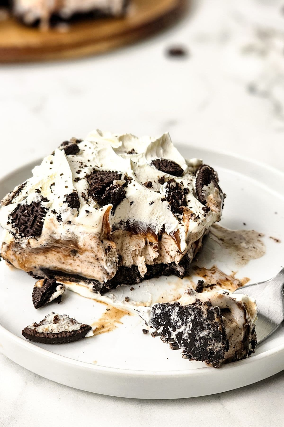 A square serving of cookies and cream dessert, reminiscent of an Oreo Ice Cream Cake, with whipped topping and cookie pieces on a white plate, a fork alongside, and a bite taken out.