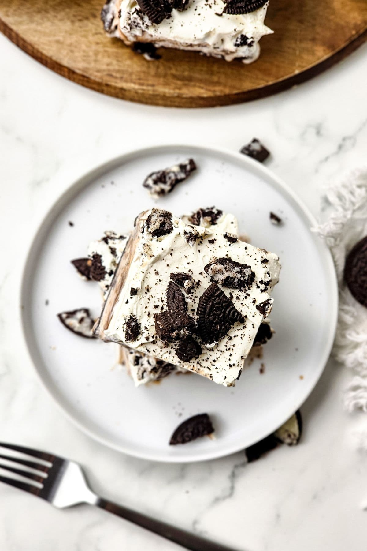 A square slice of cookies and cream dessert, reminiscent of an Oreo Ice Cream Cake, is topped with whipped cream and crumbled chocolate cookies on a white plate.