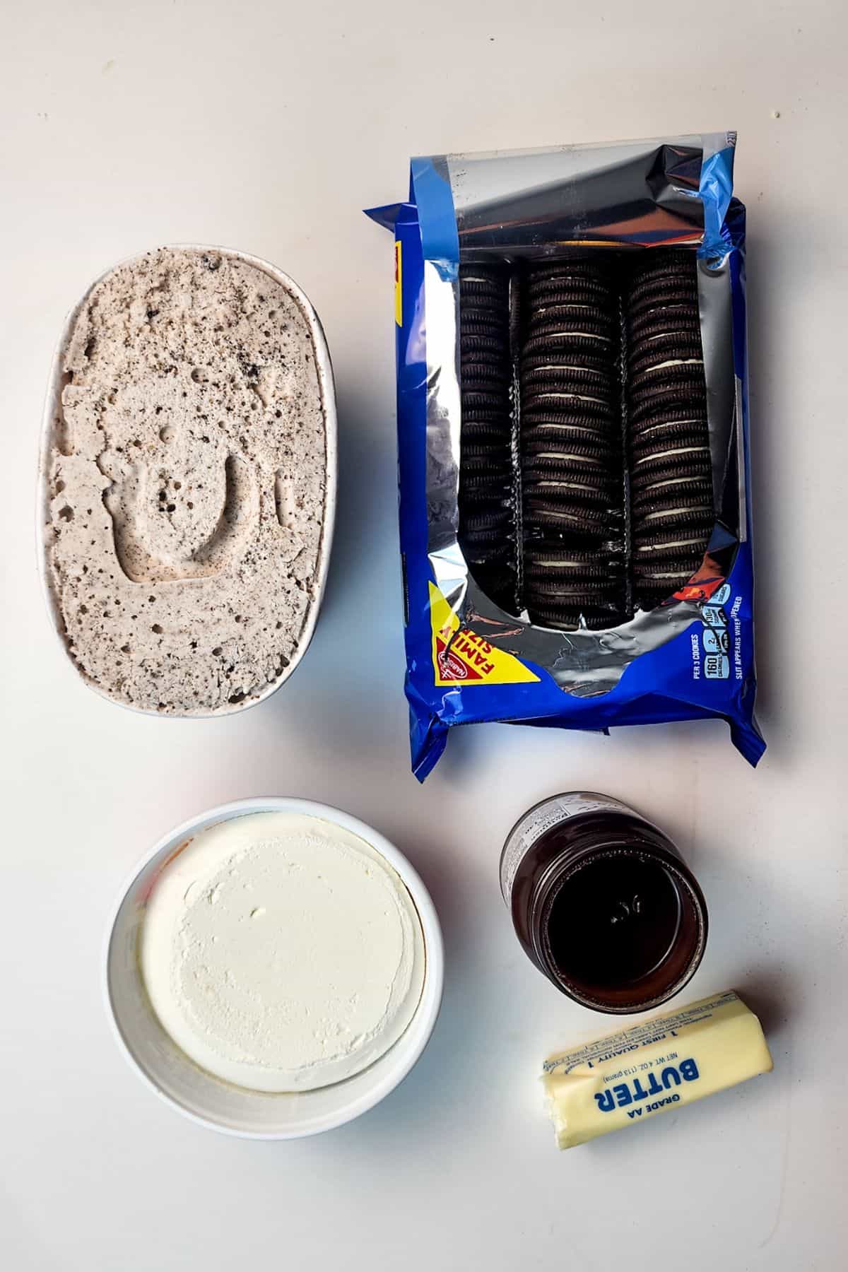 Ingredients for Oreo Ice Cream Cake beautifully arranged on a white surface, featuring ice cream with cookie pieces, a package of chocolate sandwich cookies, a bowl of cream cheese, a jar of chocolate spread, and a stick of butter.
