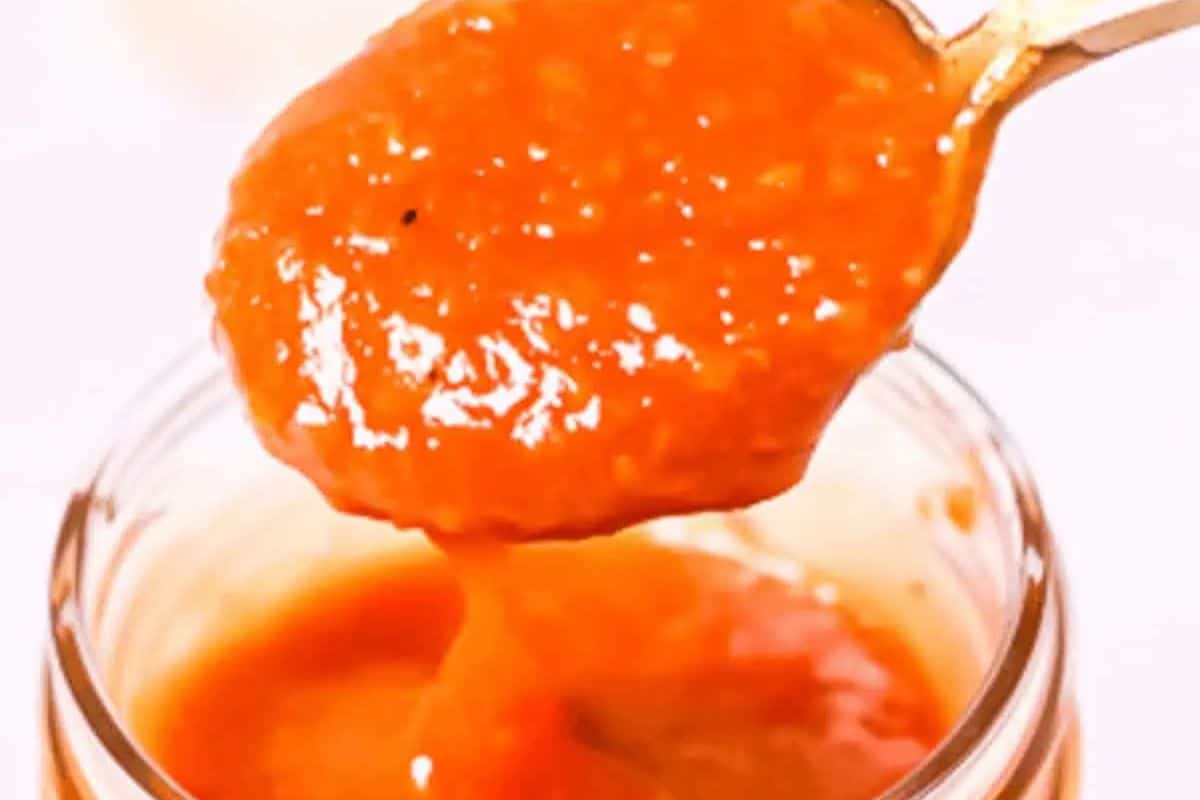A spoonful of thick, orange-red homemade BBQ sauce is being held over an open jar filled with the same sauce.
