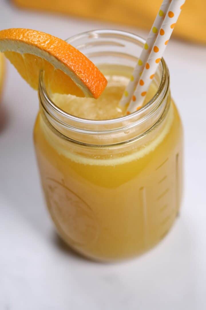 Mason jar of pineapple moonshine with an orange slice garnish and two patterned paper straws.