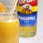 A glass of pineapple moonshine garnished with an orange slice, with a bottle of pineapple syrup in the background.