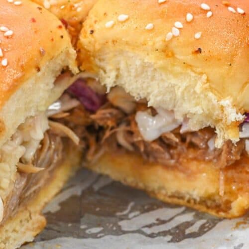 Close-up of pork sliders with coleslaw on sesame buns, featuring tender shredded pork, placed on a white surface.